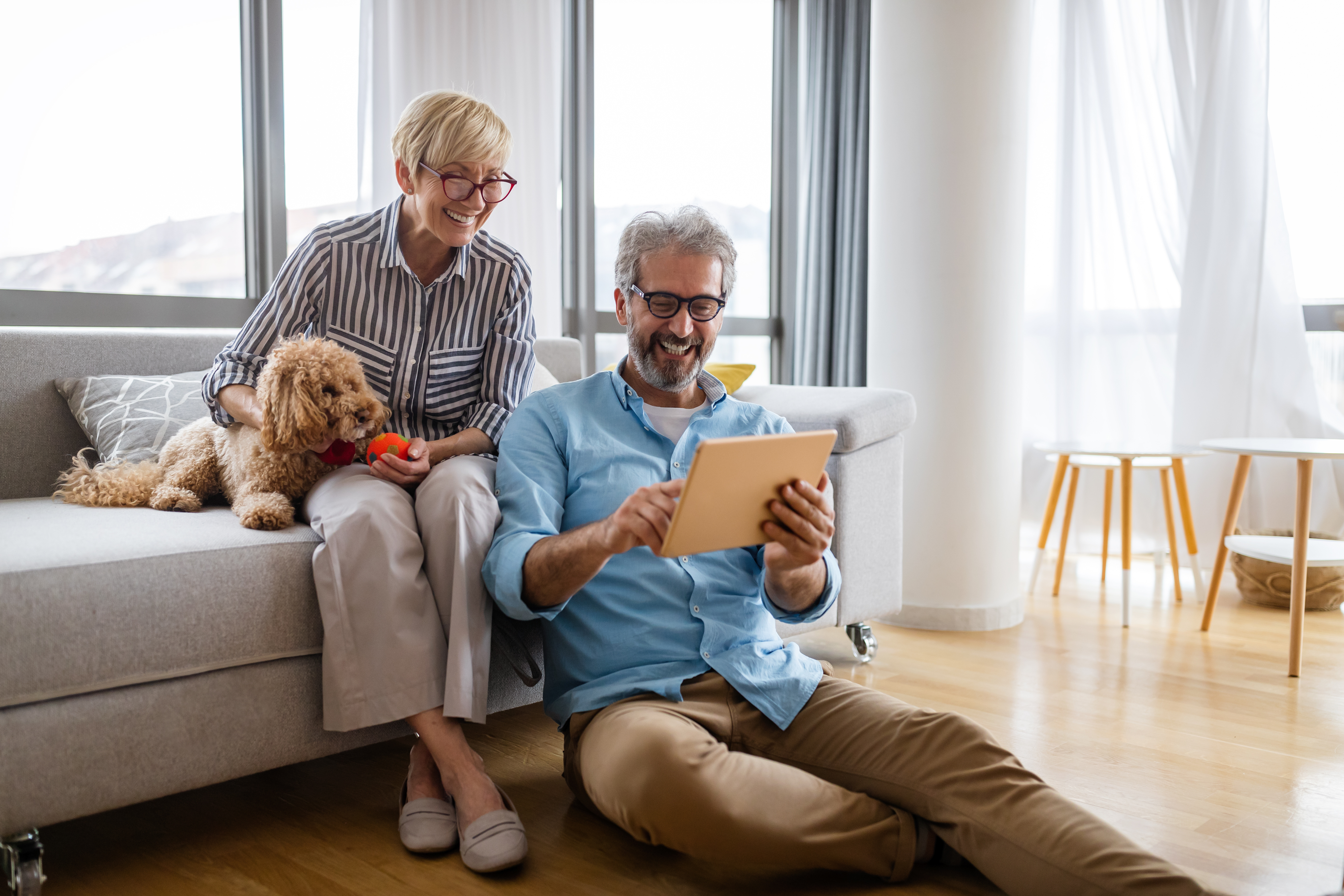 older couple sitting in living room with dog and tablet. 